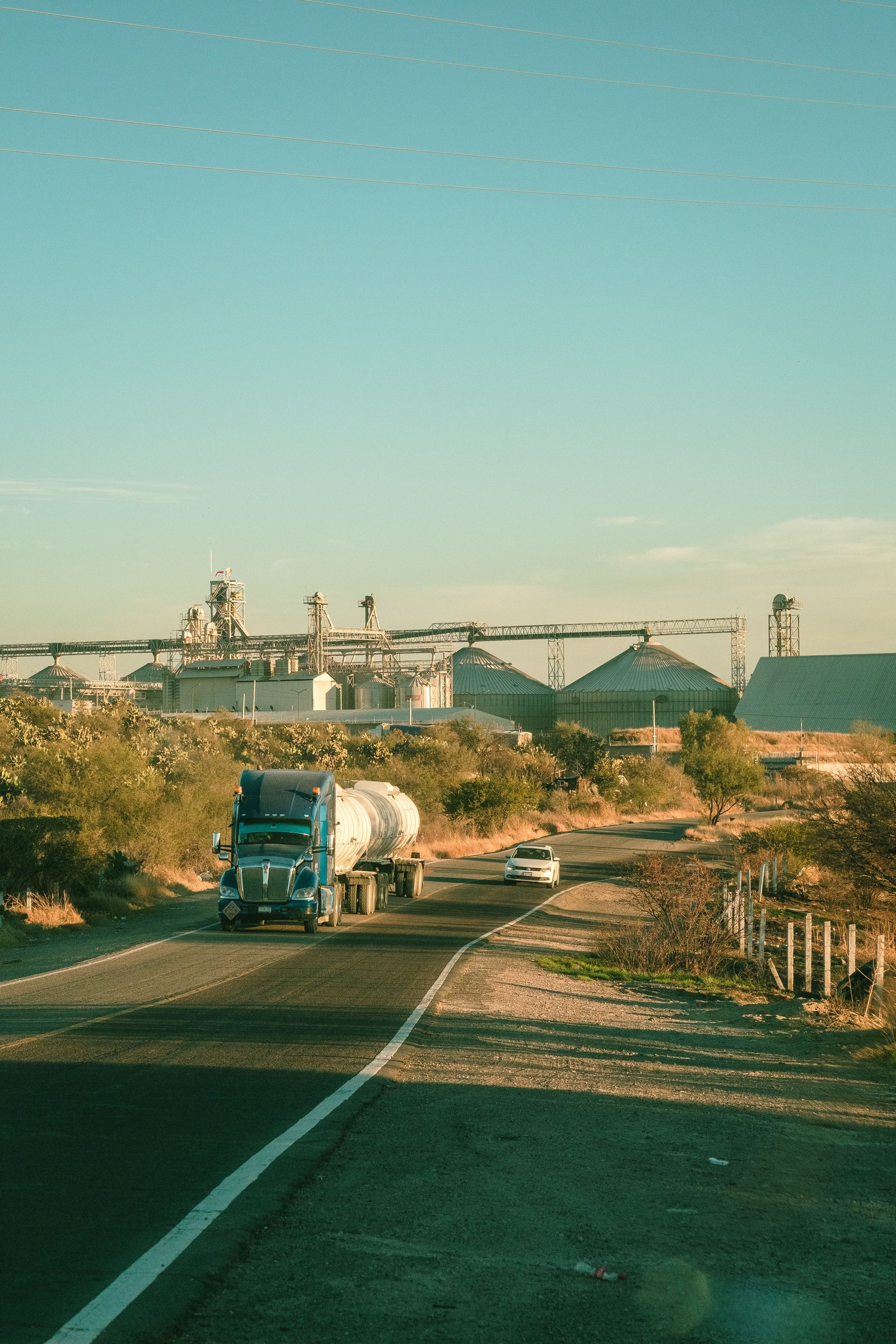 Blue conventional semi truck with a tank trailer on a road beside an industrial site